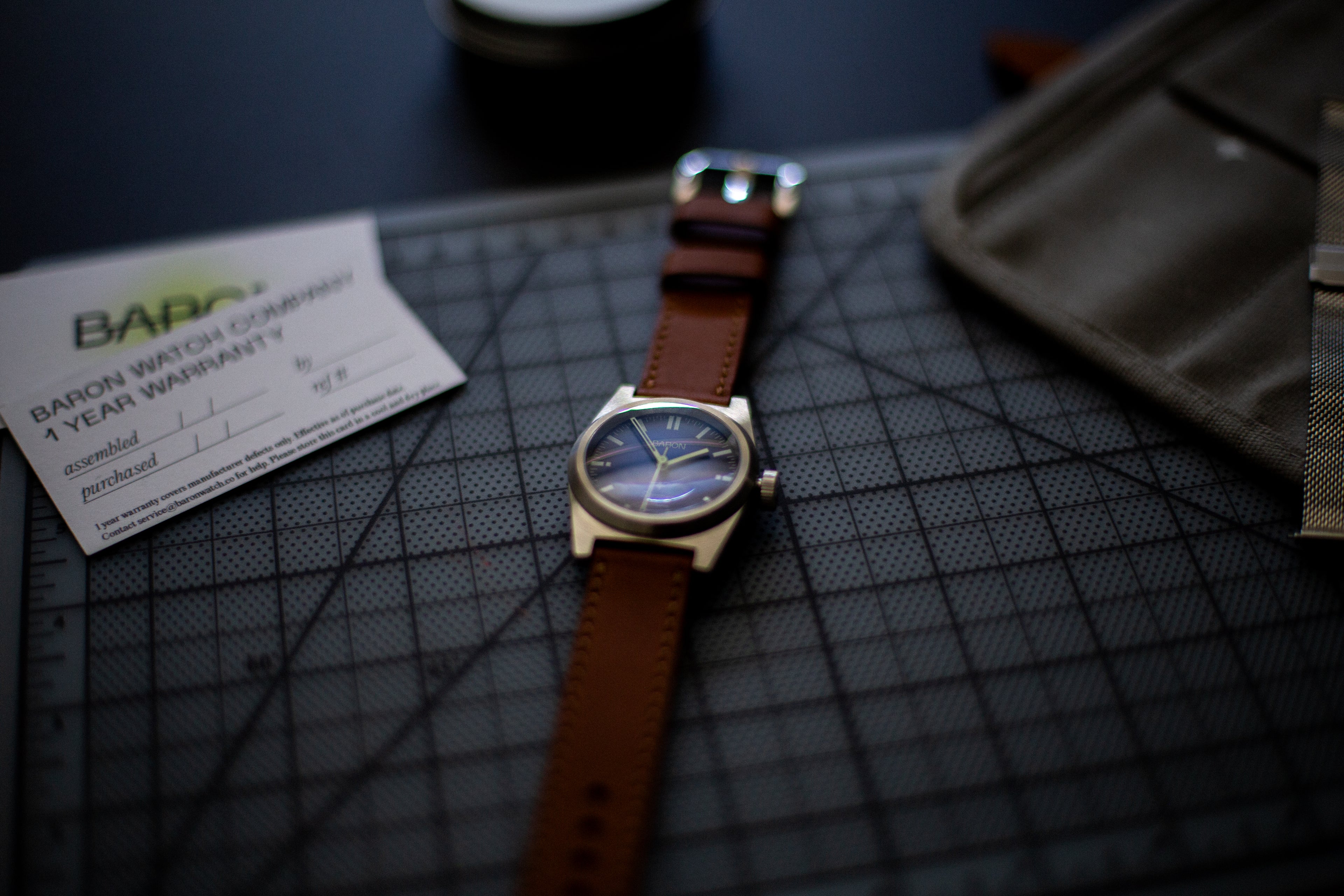 Watch with brown leather strap on a grid surface, with a card in the background.