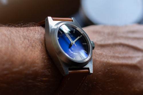 Wrist wearing a watch with a blue face and brown strap.